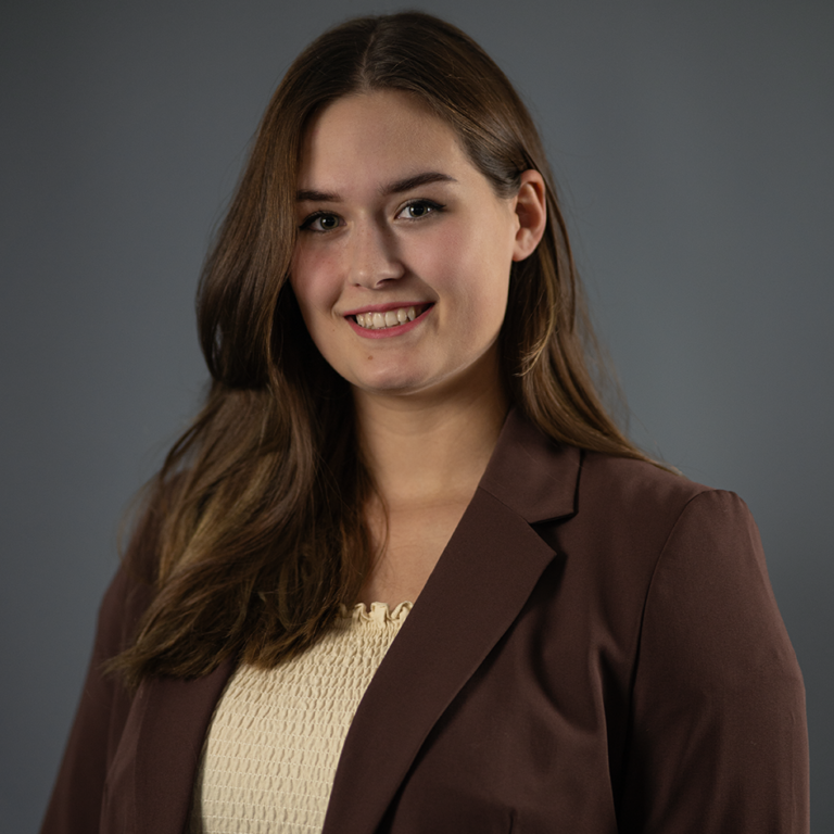 Ein Foto von Sandra Horvath. Sie lächelt in die Kamera und befindet sich vor einem dunkelgrauen Hintergrund. Sie trägt einen dunkelbraunen Blazer und ein beiges Oberteil. Ihre Haare sind brustlang, dunkelbraun und glatt.